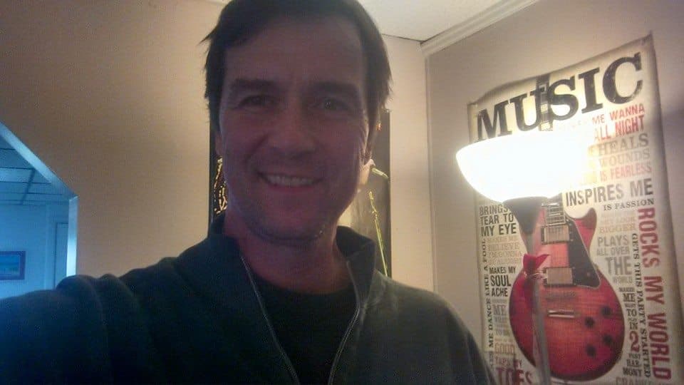 Smiling man in a dark jacket stands beside a music poster and a bright lamp.